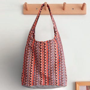 Può includere: Una borsa tote fantasia con un design geometrico in rosso, arancione, blu e nero. La borsa ha un fondo grigio e lunghe cinghie. È appesa a un attaccapanni in legno su un muro bianco.