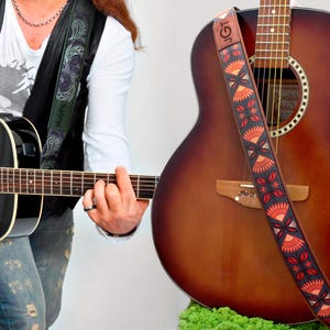 K&ouml;nnte beinhalten: Eine Person spielt eine schwarze Akustikgitarre mit einem gr&uuml;n-schwarzen Paisley-Gitarrengurt. Im Hintergrund befindet sich eine braune Akustikgitarre mit einem rot-orange-schwarzen gemusterten Gitarrengurt. Der Gurt tr&auml;gt den Text "JGT".