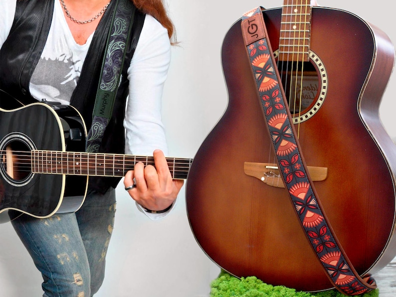 K&ouml;nnte beinhalten: Eine Person spielt eine schwarze Akustikgitarre mit einem gr&uuml;n-schwarzen Paisley-Gitarrengurt. Im Hintergrund befindet sich eine braune Akustikgitarre mit einem rot-orange-schwarzen gemusterten Gitarrengurt. Der Gurt tr&auml;gt den Text "JGT".