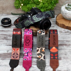 May include: Four colourful camera straps with personalised names: Adam, JGT Photography, Smith Jennifer, and Grace. Each strap features a different patterned design.