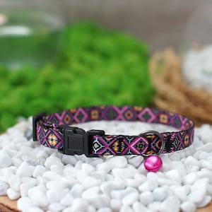 May include: A colorful patterned cat collar with a pink bell. The collar is black with pink, orange, and yellow geometric shapes.