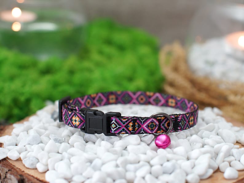 May include: A colorful patterned cat collar with a pink bell. The collar is black with pink, orange, and yellow geometric shapes.
