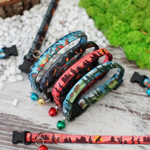 May include: Four colourful cat collars with a nature-themed print and a bell on each. The collars are stacked on top of each other, with the bottom collar being a red and orange colour with a desert scene. The second collar is a blue and green colour with a forest scene. The third collar is a black and grey colour with a mountain scene. The top collar is a blue and green colour with a forest scene. The collars are all made of a soft, comfortable material and are perfect for cats of all sizes.