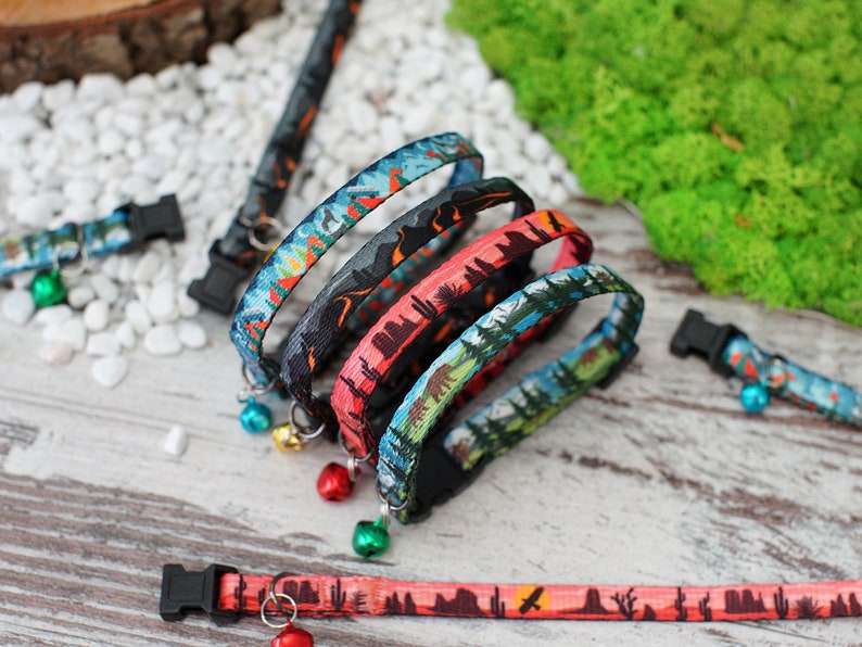May include: Four colourful cat collars with a nature-themed print and a bell on each. The collars are stacked on top of each other, with the bottom collar being a red and orange colour with a desert scene. The second collar is a blue and green colour with a forest scene. The third collar is a black and grey colour with a mountain scene. The top collar is a blue and green colour with a forest scene. The collars are all made of a soft, comfortable material and are perfect for cats of all sizes.