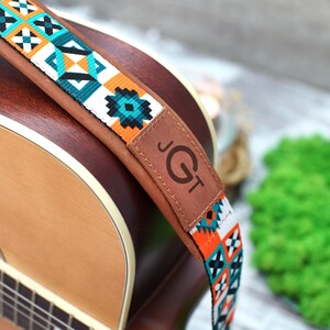 May include: A brown leather guitar strap with a colorful geometric pattern and the text "JGT" on the leather. The strap is attached to a brown acoustic guitar.