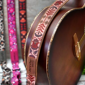 K&ouml;nnte beinhalten: Eine braune Akustikgitarre mit einem floralen Gitarrengurt. Der Gurt hat ein Leder-Etikett mit dem Text "Jennifer Smith".