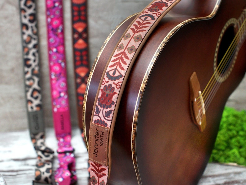 K&ouml;nnte beinhalten: Eine braune Akustikgitarre mit einem floralen Gitarrengurt. Der Gurt hat ein Leder-Etikett mit dem Text "Jennifer Smith".