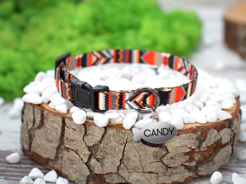 May include: A colorful cat collar with a geometric pattern in orange, black, white, and gray. The collar has a black plastic buckle and a silver metal tag that says "CANDY".