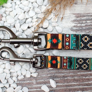 K&ouml;nnte beinhalten: Eine schwarze und gr&uuml;ne Hundeleine mit einem bunten geometrischen Muster. Die Leine hat zwei silberne Clips.