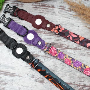 Op de afbeelding: Drie hondenhalsbanden met gepersonaliseerde naambordjes. De halsbanden zijn van zwart, paars en bordeauxrood leer met een zilveren gesp. De halsbanden zijn versierd met bloemenpatronen en een zwart en oranje camouflagepatroon.