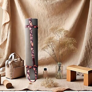 May include: A rolled grey yoga mat with a floral strap, a beige tote bag, and a small wooden bench are displayed. A glass vase with dried flowers, a small bottle, and a basket with dried flowers are also present. The items are arranged on a neutral-toned surface.