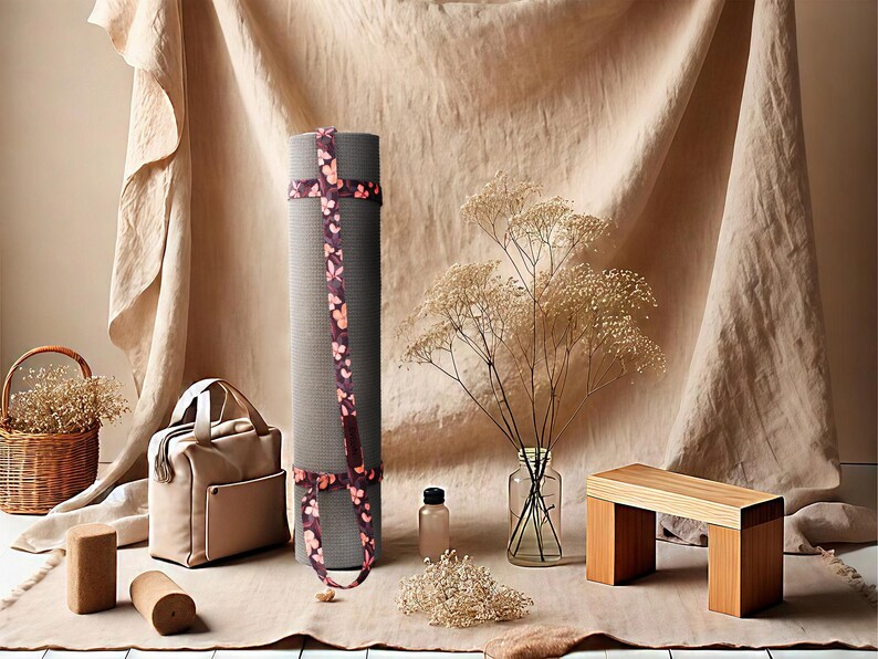 May include: A rolled grey yoga mat with a floral strap, a beige tote bag, and a small wooden bench are displayed. A glass vase with dried flowers, a small bottle, and a basket with dried flowers are also present. The items are arranged on a neutral-toned surface.