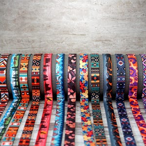 Puede incluir: Una colección de correas estampadas coloridas, probablemente para esterillas de yoga, exhibidas sobre una base cilíndrica gris. Las correas presentan varios diseños, incluyendo patrones florales, geométricos e inspirados en la naturaleza, en colores como azul, naranja y morado.