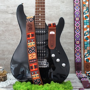 May include: A black electric guitar with a brown leather strap featuring a colorful geometric pattern. The strap has the text "Clay" on it. Three other guitar straps with different geometric patterns are visible in the background.