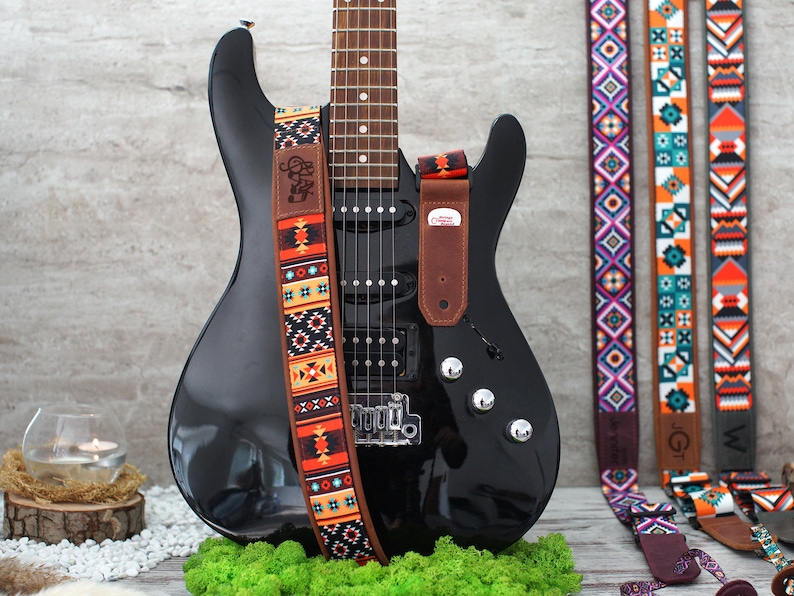 May include: A black electric guitar with a brown leather strap featuring a colorful geometric pattern. The strap has the text "Clay" on it. Three other guitar straps with different geometric patterns are visible in the background.