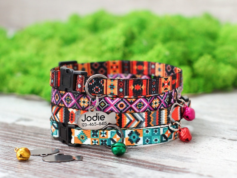 May include: A colorful cat collar with a silver tag that reads "Jodie 123-465-8415". The collar has a black buckle and is decorated with a geometric pattern in shades of orange, pink, blue, and green. The collar has multiple bells attached.