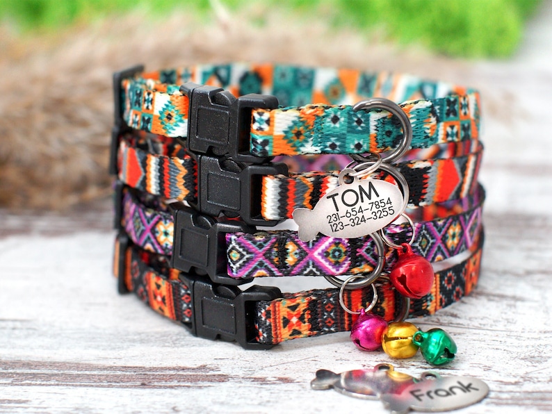 May include: Four colorful cat collars with a geometric pattern. The collars have black buckles and are stacked on top of each other. A silver tag with the name "TOM" and phone numbers is attached to the top collar. There are also silver bells attached to the collars.