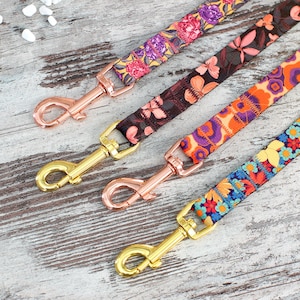 Può includere: Quattro guinzagli per cani colorati con motivi floreali e fibbie in metallo dorate e dorate rosa. I guinzagli sono su uno sfondo di legno chiaro.