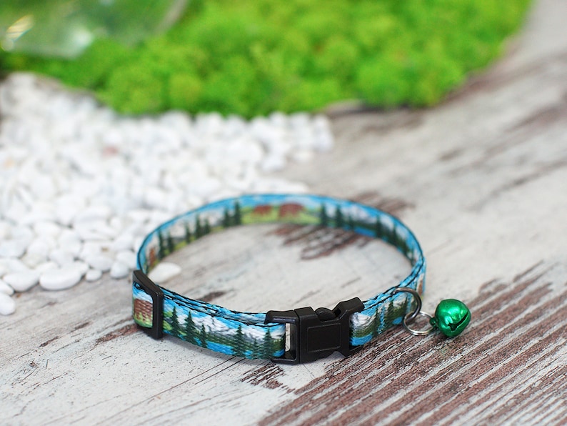 May include: A blue and green cat collar with a black buckle and a green bell. The collar features a printed design of a mountain scene.