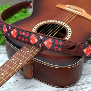 K&ouml;nnte beinhalten: Ein roter, schwarzer und wei&szlig;er Gitarrengurt mit dem aufgedruckten Wort "Grace". Der Gurt befindet sich auf einer braunen Akustikgitarre.