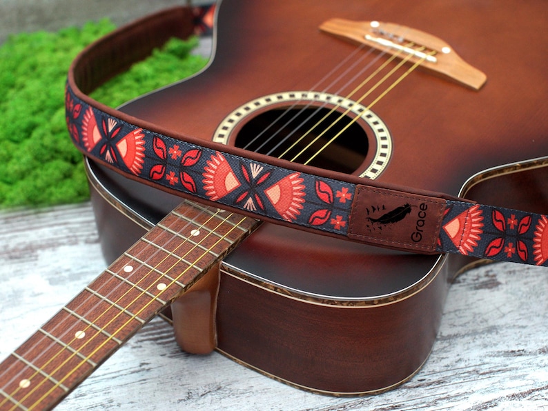 K&ouml;nnte beinhalten: Ein roter, schwarzer und wei&szlig;er Gitarrengurt mit dem aufgedruckten Wort "Grace". Der Gurt befindet sich auf einer braunen Akustikgitarre.