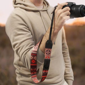 May include: A person holding a black camera with a brown and red patterned camera strap. The strap has a geometric design.