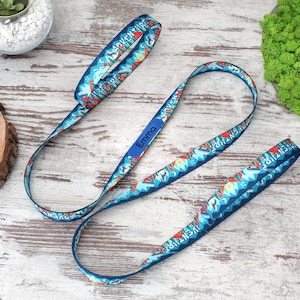 May include: Blue lanyard with a repeating mountain and arrow pattern, the word "ADVENTURE" is printed on the lanyard. The name "Emma" is printed on a blue rectangle. The lanyard is on a light wood surface.