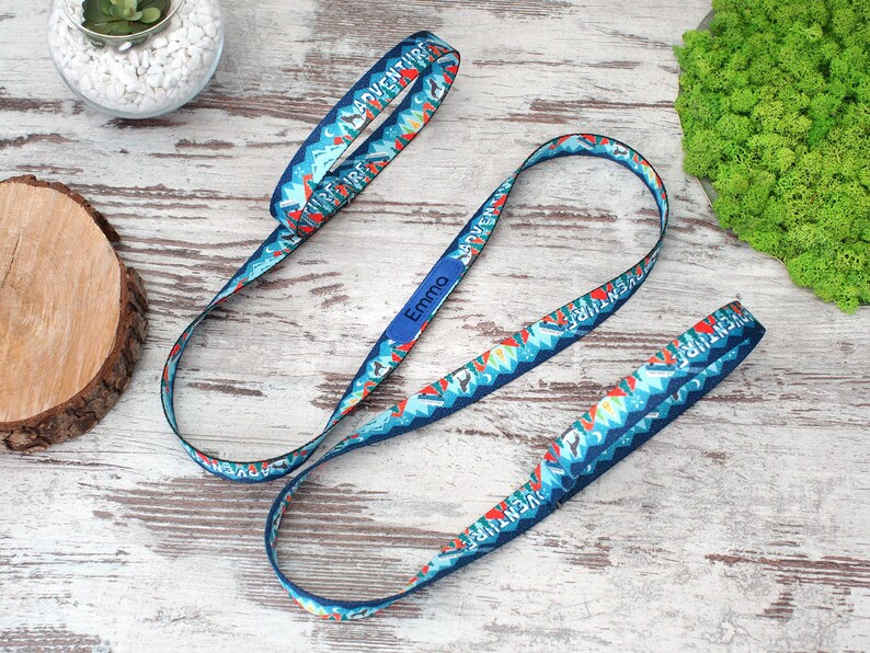 May include: Blue lanyard with a repeating mountain and arrow pattern, the word "ADVENTURE" is printed on the lanyard. The name "Emma" is printed on a blue rectangle. The lanyard is on a light wood surface.