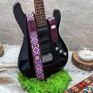 May include: A black electric guitar with a colorful patterned guitar strap. The strap has the text "Jennifer" on it. The guitar is sitting on a bed of green moss.