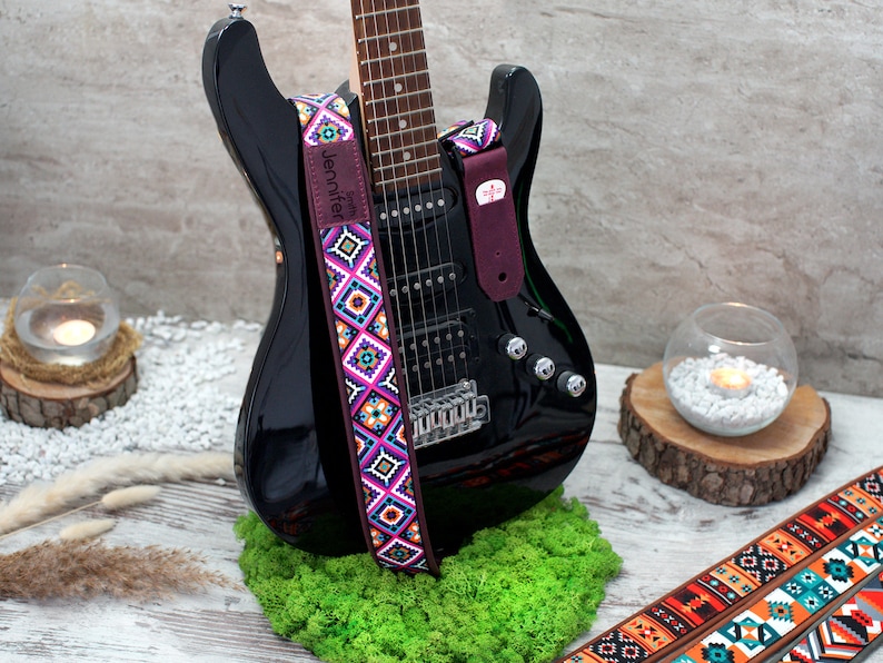 May include: A black electric guitar with a colorful patterned guitar strap. The strap has the text "Jennifer" on it. The guitar is sitting on a bed of green moss.