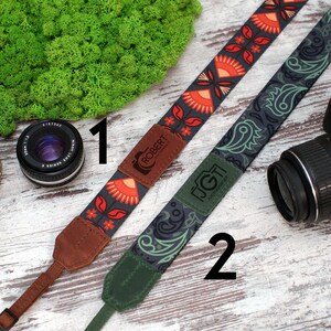 May include: Two camera straps with different patterns. The first strap is black with red and orange floral designs and the text "ROBERT". The second strap is green with black paisley designs and the text "JGT".