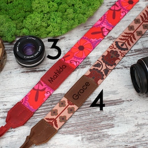 May include: Two camera straps with different patterns. One strap is red, pink and white with the name "Matilda" printed on it. The other strap is brown, beige and red with the name "Grace" printed on it.