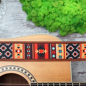 May include: A brown leather guitar strap with a colorful geometric pattern in shades of red, orange, black, white, and blue. The strap is attached to an acoustic guitar.