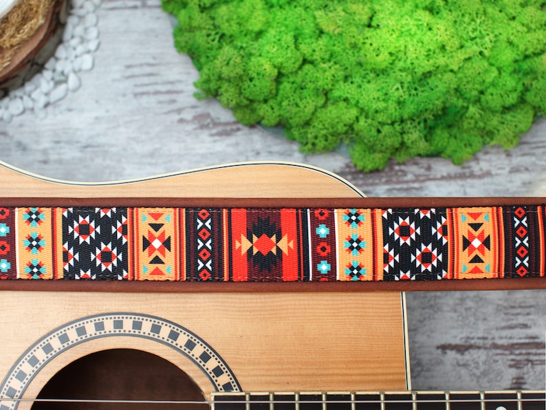 May include: A brown leather guitar strap with a colorful geometric pattern in shades of red, orange, black, white, and blue. The strap is attached to an acoustic guitar.