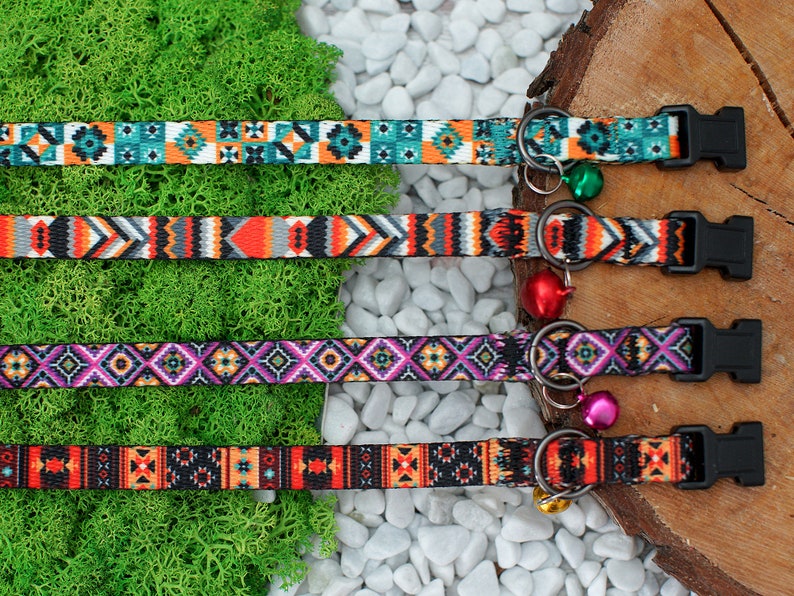 May include: Four colorful patterned cat collars with black buckles and bell charms. The collars feature geometric designs in shades of orange, teal, purple, and black.