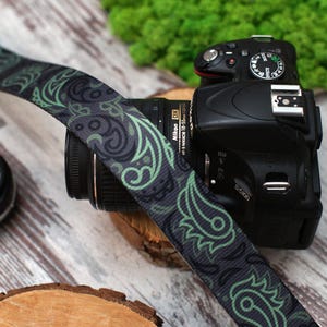 May include: A black camera with a green and black patterned camera strap. The camera has a lens attached and a black flash on top.