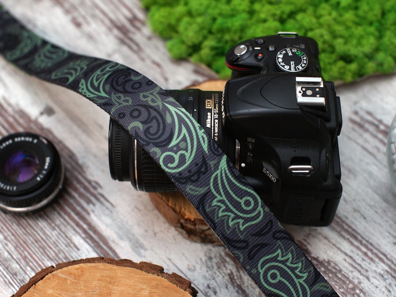 May include: A black camera with a green and black patterned camera strap. The camera has a lens attached and a black flash on top.