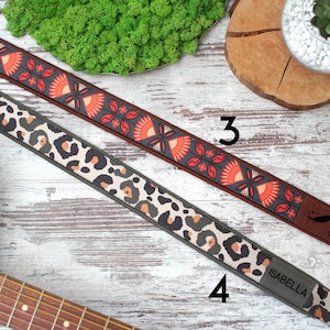 K&ouml;nnte beinhalten: Zwei Gitarrengurte mit unterschiedlichen Mustern. Ein Gurt hat ein rotes, oranges und schwarzes Blumenmuster mit dem Text "Groove" am Ende. Der andere Gurt hat ein schwarzes, wei&szlig;es und braunes Leopardenmuster mit dem Text "Isabella" am Ende.