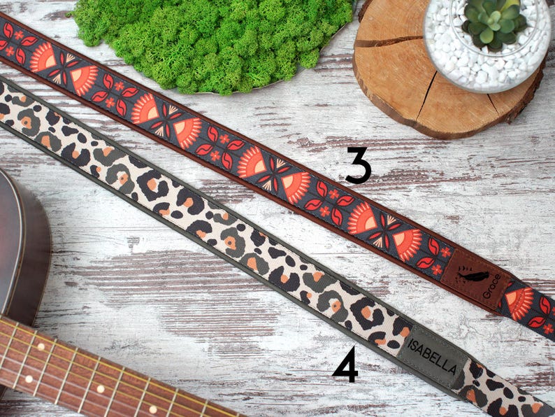K&ouml;nnte beinhalten: Zwei Gitarrengurte mit unterschiedlichen Mustern. Ein Gurt hat ein rotes, oranges und schwarzes Blumenmuster mit dem Text "Groove" am Ende. Der andere Gurt hat ein schwarzes, wei&szlig;es und braunes Leopardenmuster mit dem Text "Isabella" am Ende.