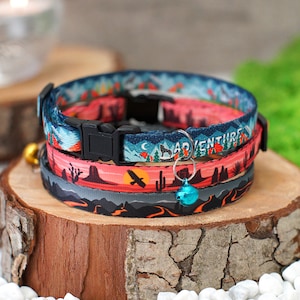 May include: Three colourful cat collars with a desert landscape, mountain, and night sky design. The collars have a black buckle and a bell charm. The text "Adventure" is printed on one of the collars.