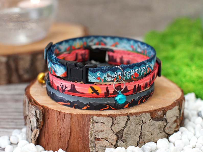 May include: Three colourful cat collars with a desert landscape, mountain, and night sky design. The collars have a black buckle and a bell charm. The text "Adventure" is printed on one of the collars.