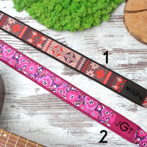 K&ouml;nnte beinhalten: Zwei Gitarrengurte mit unterschiedlichen Mustern. Der erste Gurt hat ein geometrisches Muster in Rot-, Braun- und Schwarzt&ouml;nen. Der zweite Gurt hat ein Blumenmuster in Rosa-, Lila- und Wei&szlig;t&ouml;nen. Beide Gurte haben schwarze Lederenden mit den Markennamen "Murphy" und "JGT" darauf gedruckt.