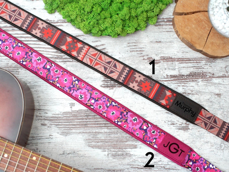 K&ouml;nnte beinhalten: Zwei Gitarrengurte mit unterschiedlichen Mustern. Der erste Gurt hat ein geometrisches Muster in Rot-, Braun- und Schwarzt&ouml;nen. Der zweite Gurt hat ein Blumenmuster in Rosa-, Lila- und Wei&szlig;t&ouml;nen. Beide Gurte haben schwarze Lederenden mit den Markennamen "Murphy" und "JGT" darauf gedruckt.