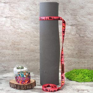 May include: A rolled grey yoga mat secured with a red strap with a desert landscape design. The strap is wrapped around the mat and has the name "Claire" printed on it. Additional patterned straps and a small succulent are also visible.