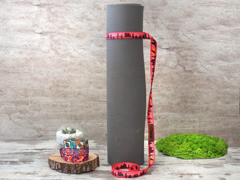 May include: A rolled grey yoga mat secured with a red strap with a desert landscape design. The strap is wrapped around the mat and has the name "Claire" printed on it. Additional patterned straps and a small succulent are also visible.