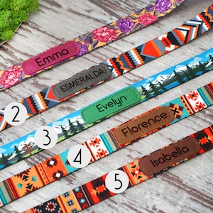 May include: A selection of colorful dog collars with personalized name tags. The collars feature various patterns, including floral, geometric, and landscape designs. Each collar has a name tag with a different name: Emma, Esmeralda, Evelyn, Florence, and Isabella.