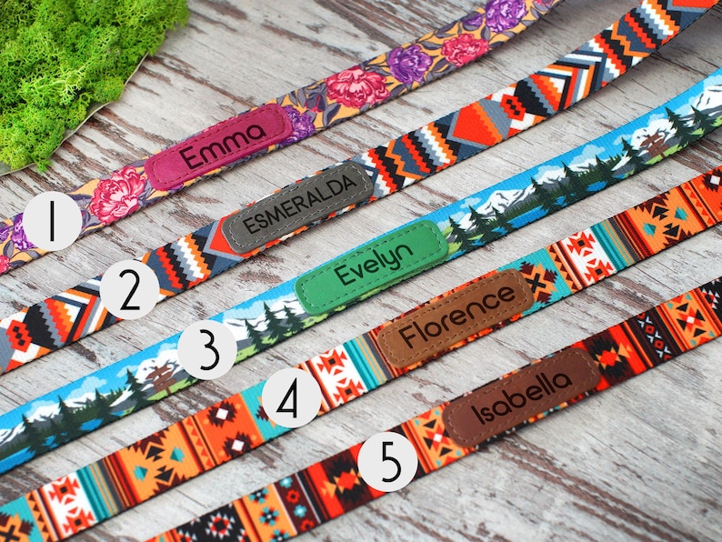 May include: A selection of colorful dog collars with personalized name tags. The collars feature various patterns, including floral, geometric, and landscape designs. Each collar has a name tag with a different name: Emma, Esmeralda, Evelyn, Florence, and Isabella.