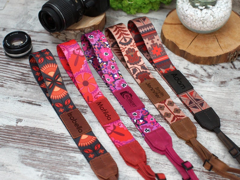 May include: Five colourful camera straps with personalised names, including Isabella, Matilda, Jennifer Smith, Adam, and Robert. The straps feature a variety of floral and geometric patterns in shades of red, pink, and brown.