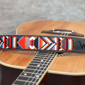 May include: A guitar strap with a colorful geometric pattern and a gray leather patch with the letter "W" on it. The strap is resting on an acoustic guitar.