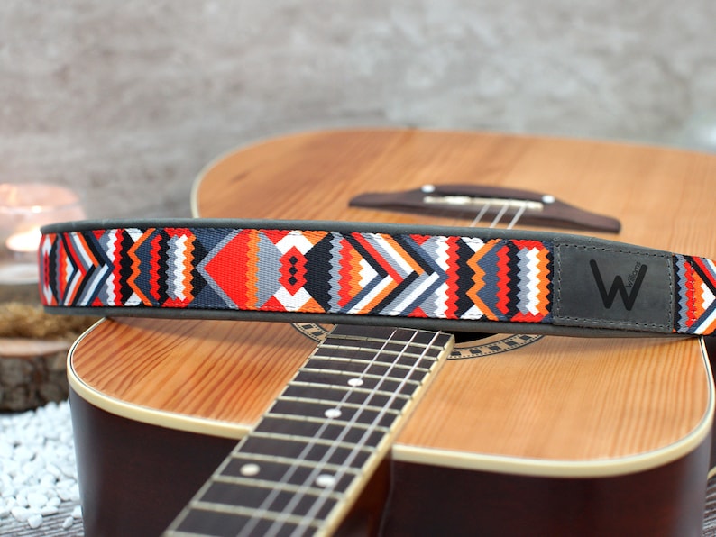 May include: A guitar strap with a colorful geometric pattern and a gray leather patch with the letter "W" on it. The strap is resting on an acoustic guitar.